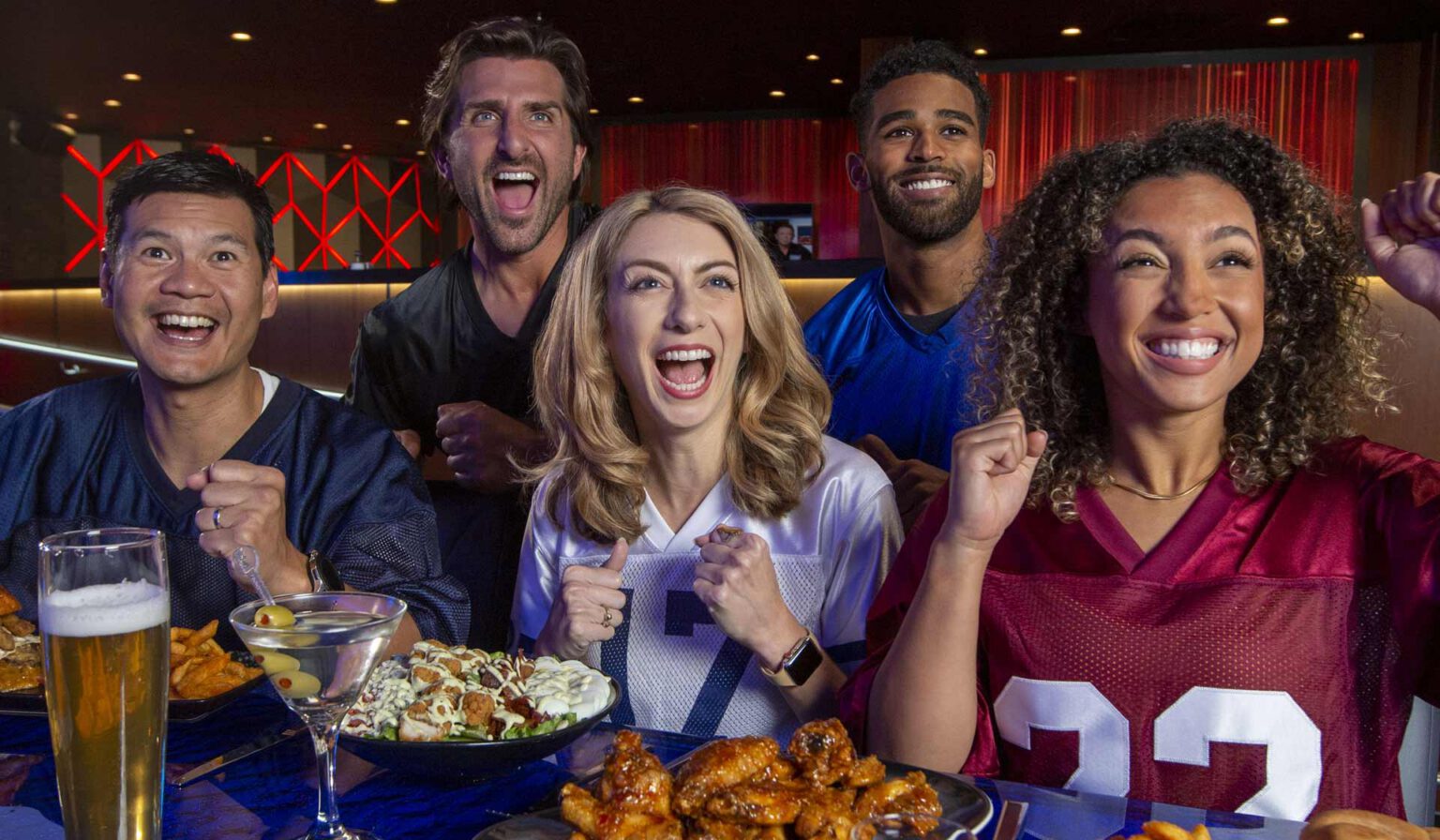 A group of people celebrating and cheering while having a salad, wings and drinks in front of themselves.