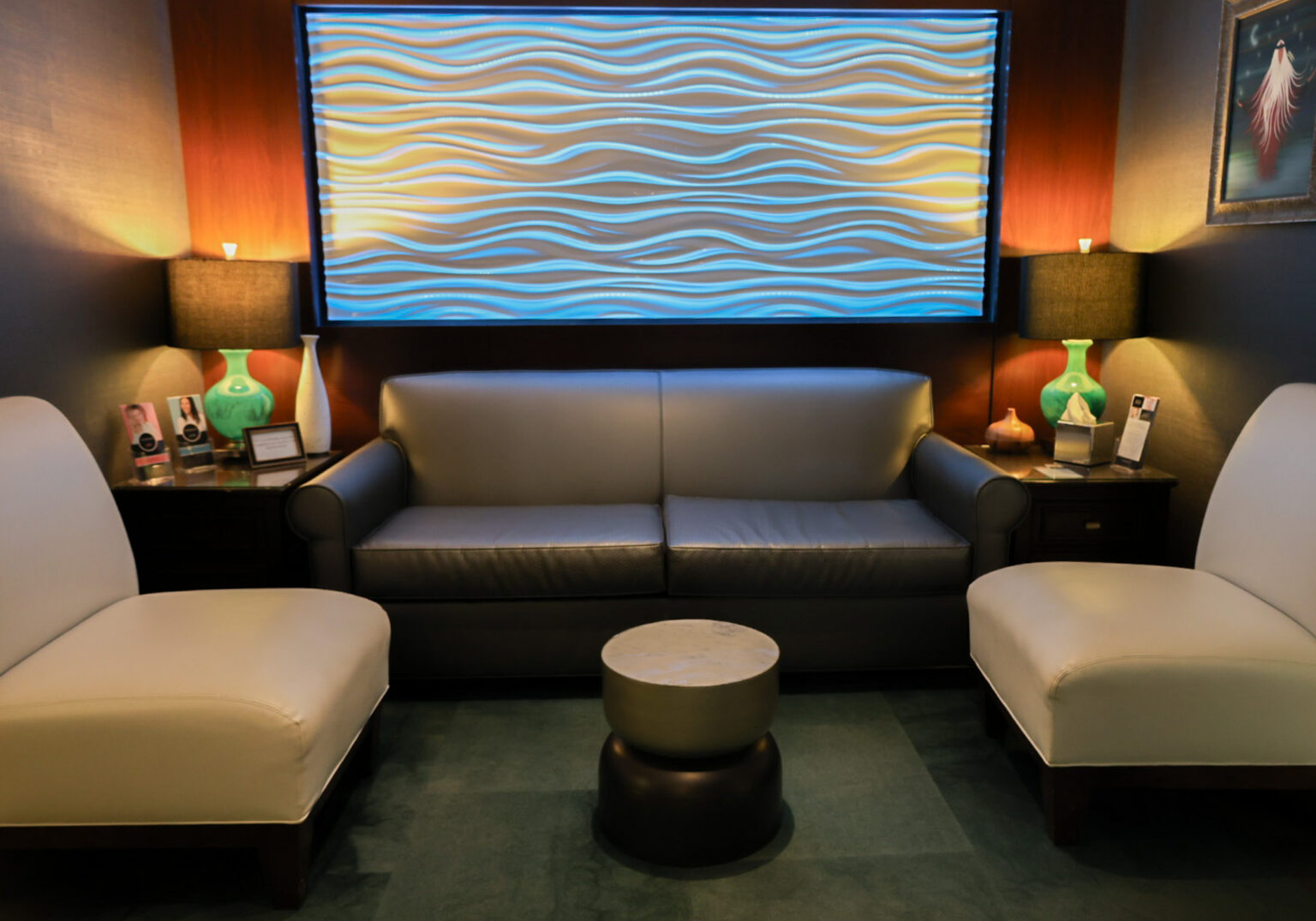 A modern seating area featuring a gray leather sofa flanked by two white armchairs. In the background, a blue and beige wave-patterned wall panel is illuminated. Two green lamps are on side tables beside the sofa, and a small round table sits in the center.