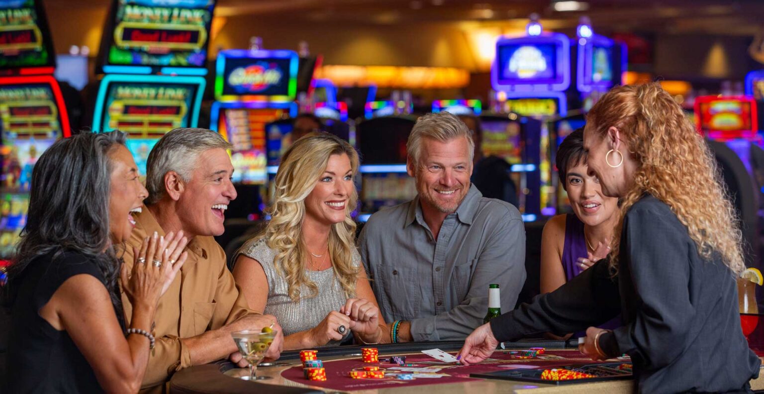 A group of five adults sitting around a casino table, interacting with a female dealer. The table has dice and poker chips, with colorful slot machines visible in the background. The individuals appear engaged and are smiling at one another.