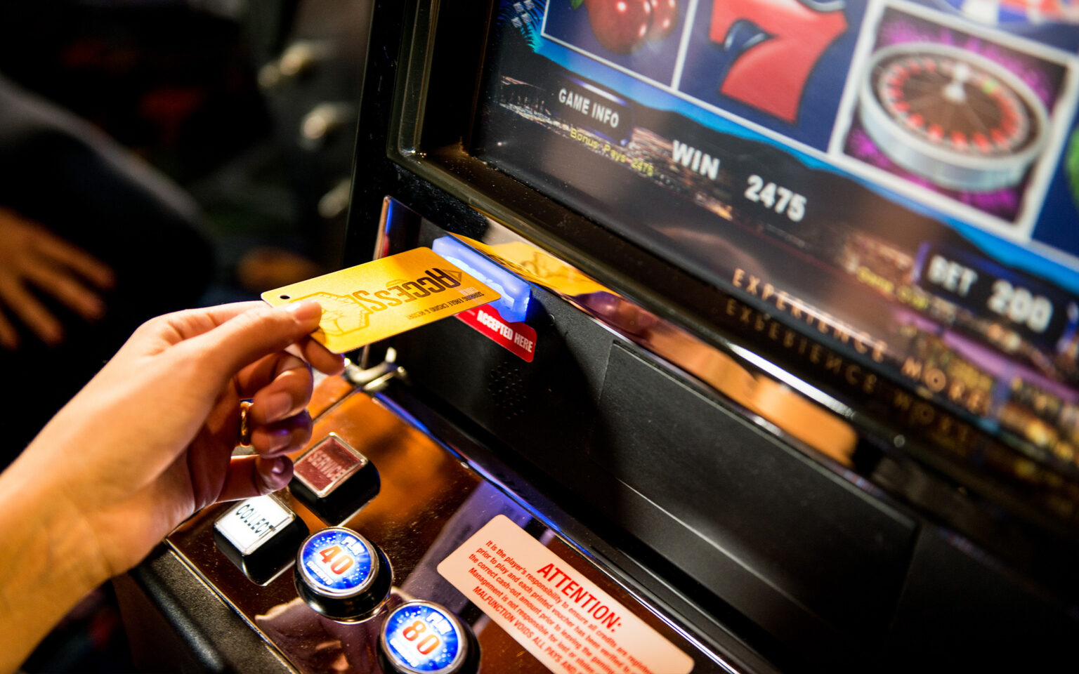 A hand inserting a yellow player card into a slot on a slot machine, with the display showing a win amount of 2475. The machine has various buttons and a sign indicating attention is needed. The background is blurred, hinting at a casino environment.