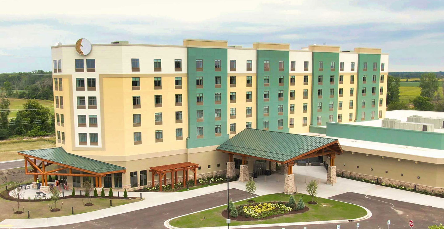 A modern hotel building featuring a combination of yellow and green facades. The entrance has a wooden canopy, landscaped gardens, and a spacious parking area. The hotel has multiple floors and large windows, set against a backdrop of open fields.