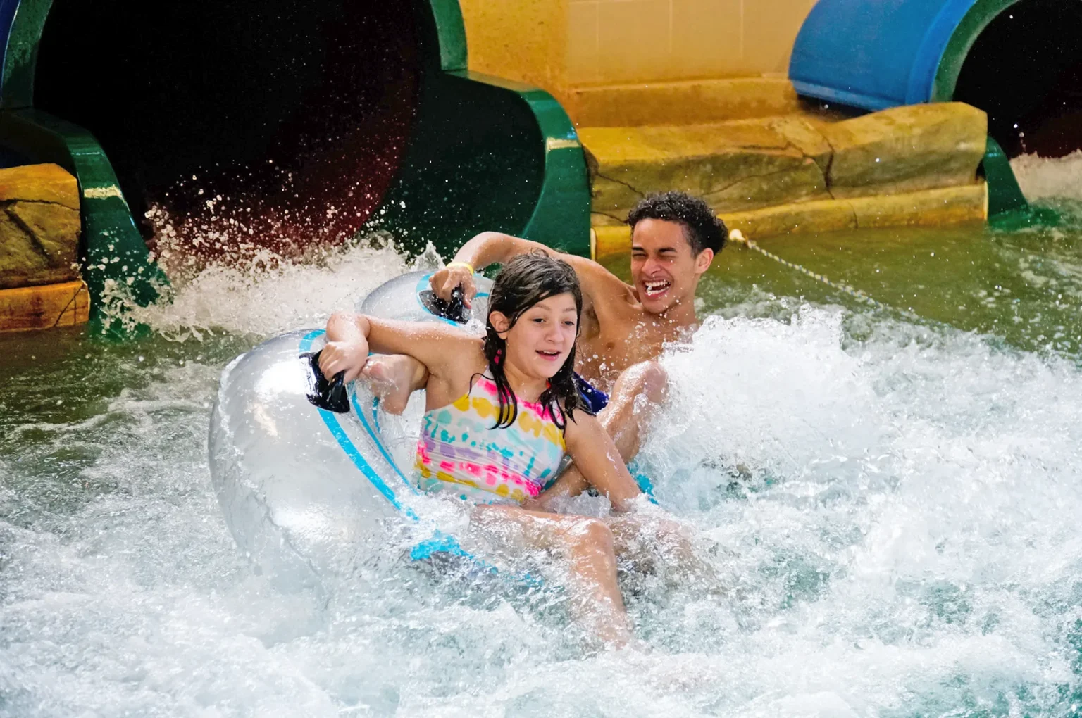 Two kids sliding down a water slide with a hige splash and both are smiling.